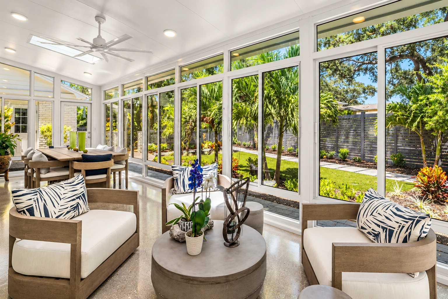 Bright four-season sunroom with white-framed windows, pale lounge chairs, and a view of a lush garden.