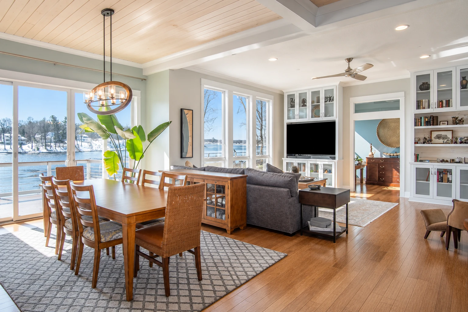 Bright open living and dining room with large windows overlooking a lake, wood flooring, a dining table, and a sofa.