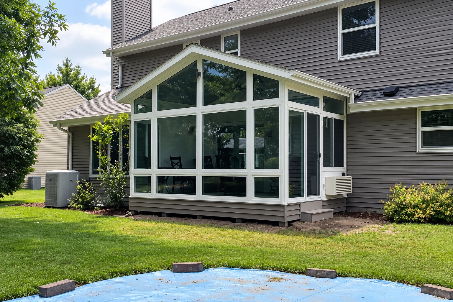 White-framed sunroom addition with a peaked roof and broad windows, set against a gray-sided house and lawn.