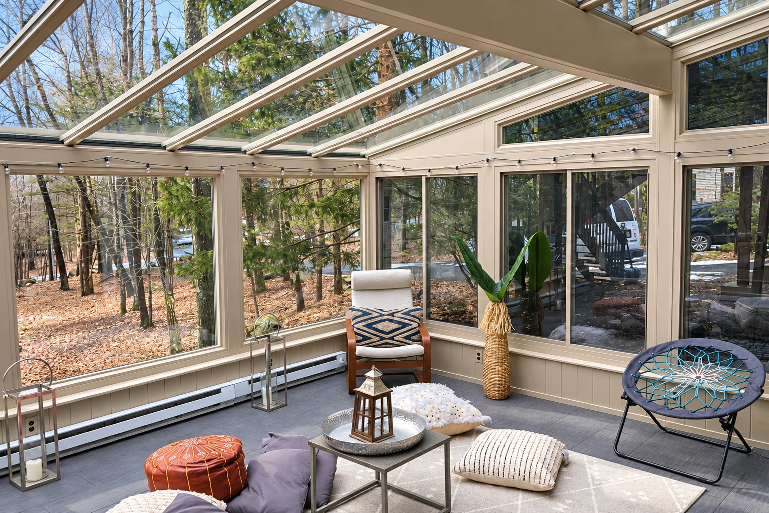 A furnished solarium has glass walls and a sloped glass roof overlooking trees, with a chair, potted plant and floor cushions.