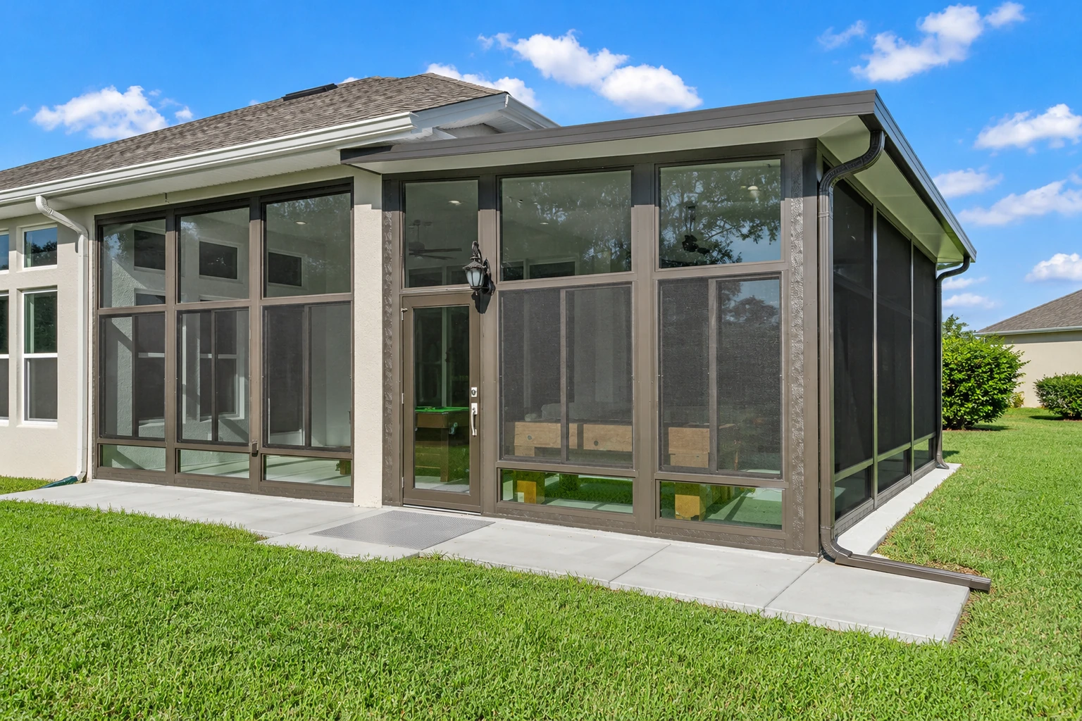 A brown-framed sunroom addition with screened windows and a glass door sits beside a white house on a sunny lawn.