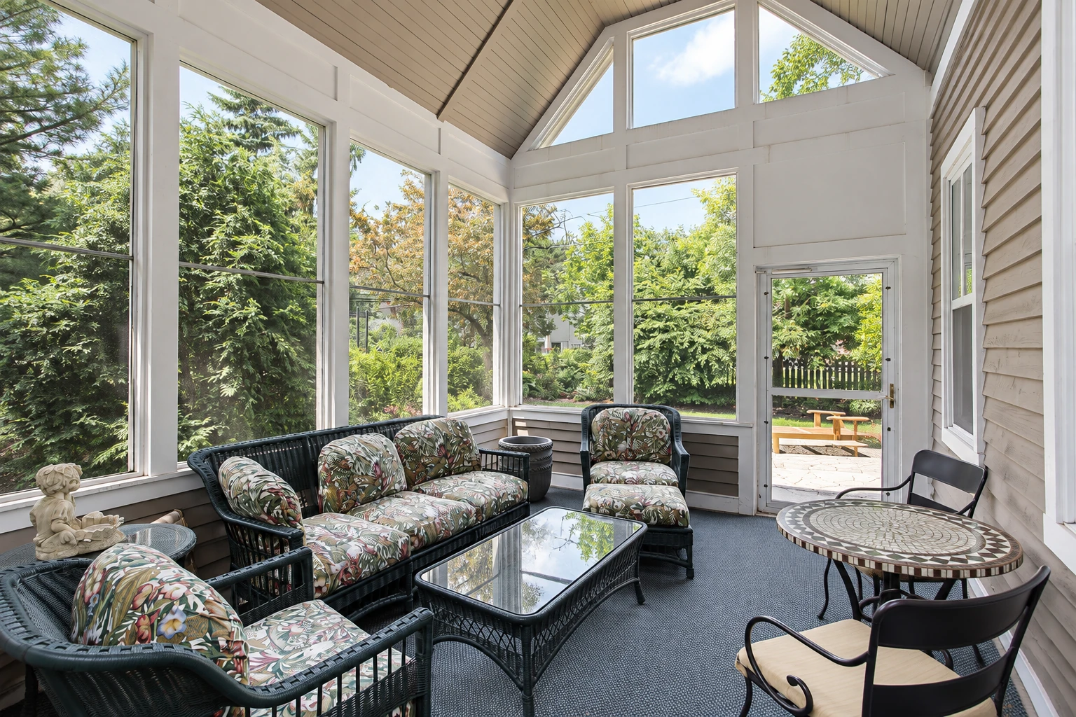 Three-season sunroom with a peaked paneled ceiling, screened windows and wicker seating around glass and mosaic tables.