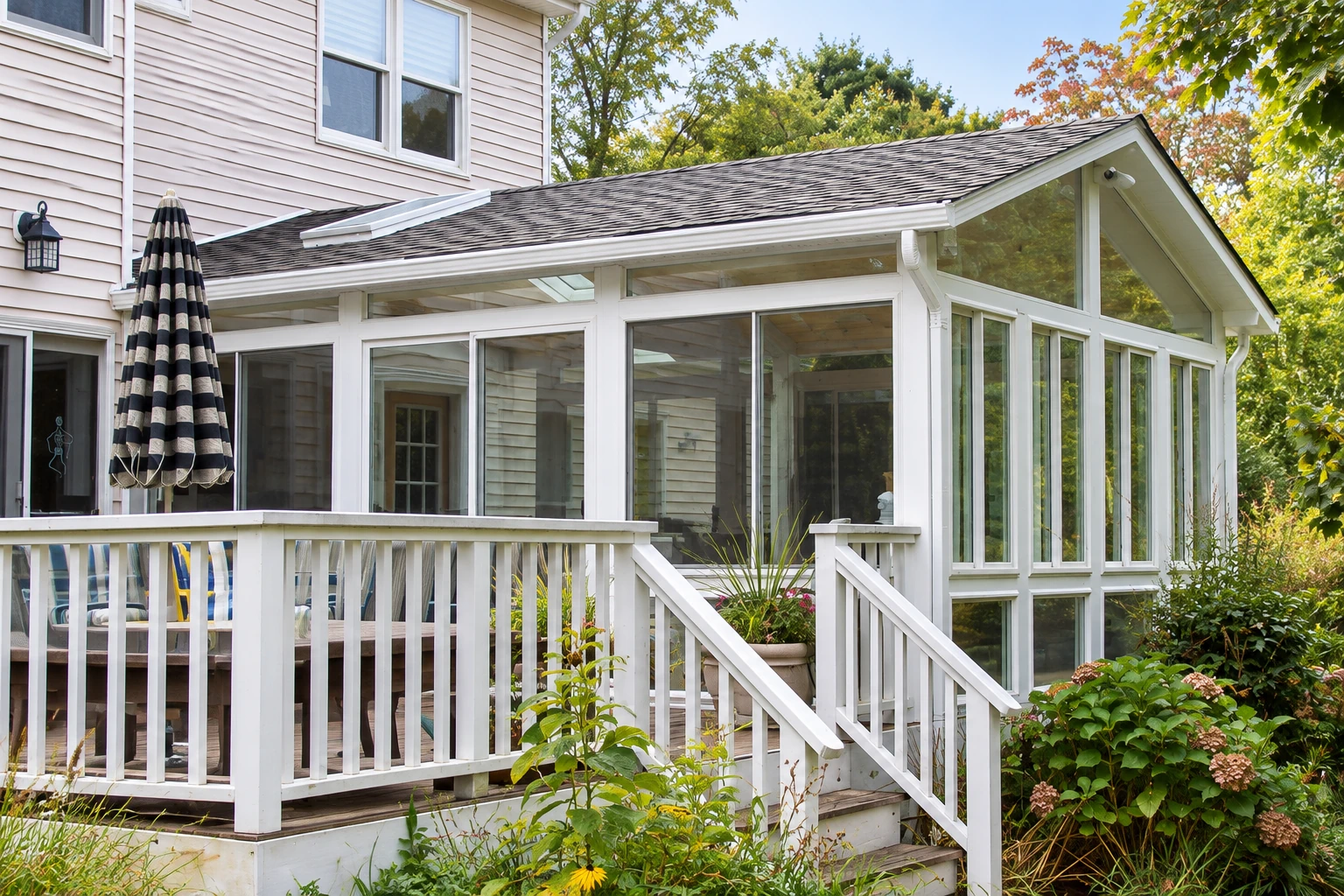 A white-framed three-season sunroom extends from a light-sided house beside a deck with white railings and garden plants.